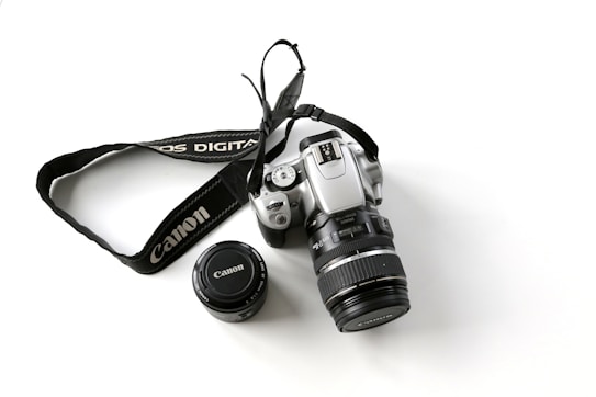 A silver digital camera with a detachable lens cap and a branded camera strap laid out on a white surface. The camera has various buttons and dials visible, suggesting a professional model.