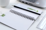 An open spiral notebook with blank pages, accompanied by a black pencil on top. On the right side, there is a silver laptop partially visible. A ruler is placed next to the notebook, along with a green paperclip and a green pushpin. A white mug is on the left, along with a green binder clip.
