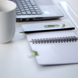 black pencil on ruled notepad beside white ceramic mug and gray laptop computer