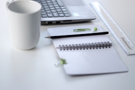 A neatly arranged workspace features an open laptop, a white coffee mug, and a spiral notebook with pages open. A black pen, green paperclip, and a small green pushpin rest on the notebook. A transparent plastic ruler with measurement markings lies beside the notebook, and the overall setting is on a clean, white surface.