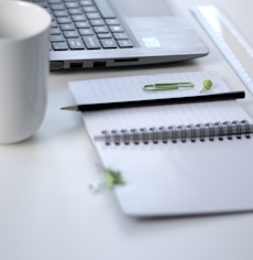 black pencil on ruled notepad beside white ceramic mug and gray laptop computer