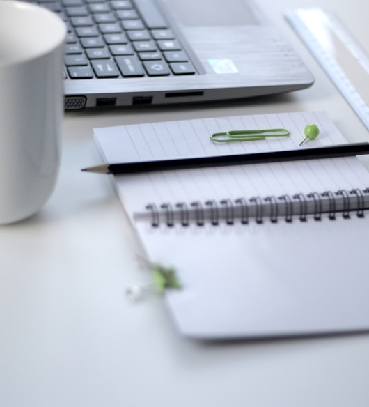 black pencil on ruled notepad beside white ceramic mug and gray laptop computer