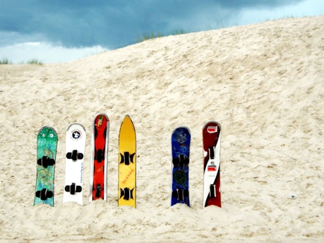 Six colorful sandboards are neatly placed upright in the sand against the backdrop of a sandy dune. The sky above is cloudy, creating a contrasting atmosphere with the bright colors of the boards.
