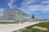 A large industrial building with the SpaceX logo and an American flag on its side. In the background, a launch pad and a water tower are visible. The foreground includes a grassy area and a paved path, with a clear blue sky above.