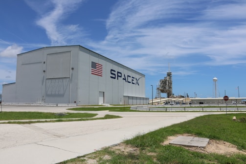 A large industrial building with the SpaceX logo and an American flag on its side. In the background, a launch pad and a water tower are visible. The foreground includes a grassy area and a paved path, with a clear blue sky above.