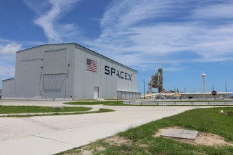 A large industrial building with the SpaceX logo and an American flag on its side. In the background, a launch pad and a water tower are visible. The foreground includes a grassy area and a paved path, with a clear blue sky above.