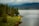 A serene lakeside scene features a small wooden cabin nestled among dense evergreen trees. A lush, grassy shoreline leads to a calm body of water extending into the distance. Mountains rise under a cloudy sky, creating a tranquil and secluded atmosphere.