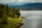 A serene lakeside scene features a small wooden cabin nestled among dense evergreen trees. A lush, grassy shoreline leads to a calm body of water extending into the distance. Mountains rise under a cloudy sky, creating a tranquil and secluded atmosphere.