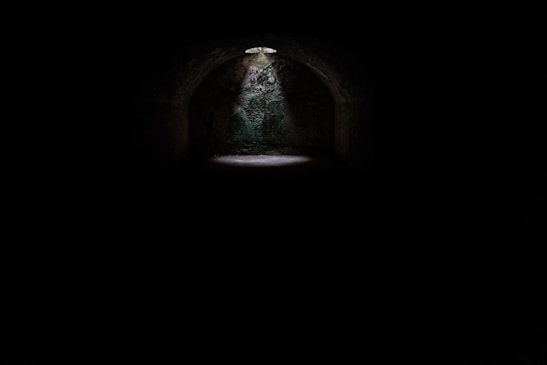 A dimly lit underground space with a stone arch and a small patch of light coming from above, illuminating the ground beneath.