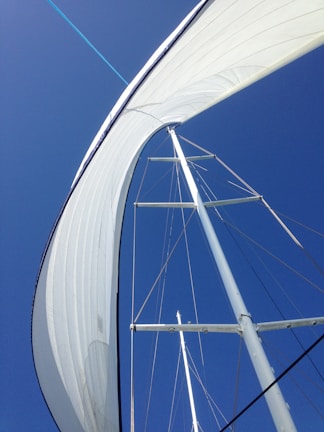 A tall sail on a sailboat is captured from below against a vivid blue sky, giving a sense of upward motion and expansive space. The sail is fully extended, showcasing its large size and elegant curve as it catches the wind. The mast and rigging are visible, adding structure and lines to the composition.
