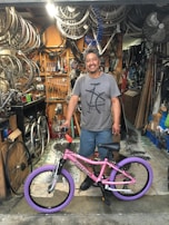 A man is standing in a bicycle repair shop, smiling with a pink bicycle featuring purple tires. The shop is cluttered with bicycle parts, including wheels and tools hanging on a pegboard, and various items around the workspace.