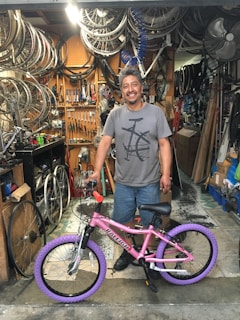A man is standing in a bicycle repair shop, smiling with a pink bicycle featuring purple tires. The shop is cluttered with bicycle parts, including wheels and tools hanging on a pegboard, and various items around the workspace.