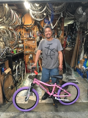 A man is standing in a bicycle repair shop, smiling with a pink bicycle featuring purple tires. The shop is cluttered with bicycle parts, including wheels and tools hanging on a pegboard, and various items around the workspace.