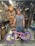 A man is standing in a bicycle repair shop, smiling with a pink bicycle featuring purple tires. The shop is cluttered with bicycle parts, including wheels and tools hanging on a pegboard, and various items around the workspace.