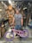 A man is standing in a bicycle repair shop, smiling with a pink bicycle featuring purple tires. The shop is cluttered with bicycle parts, including wheels and tools hanging on a pegboard, and various items around the workspace.