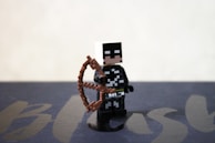 A LEGO-style figurine is holding a brown bow, dressed in a black and white costume. The background is simple and blurred, with a faint suggestion of text or design underneath.