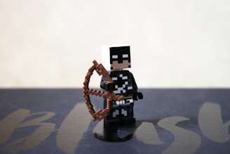 A LEGO-style figurine is holding a brown bow, dressed in a black and white costume. The background is simple and blurred, with a faint suggestion of text or design underneath.
