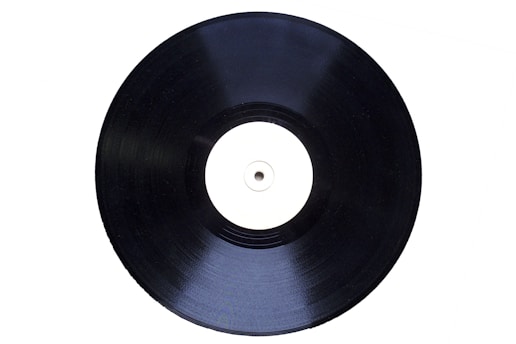 A black vinyl record with a plain white label in the center is displayed at a slight angle against a white background.