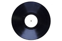 A black vinyl record with a plain white label in the center is displayed at a slight angle against a white background.