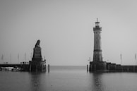 A black and white image features a waterfront scene, including a prominent lighthouse on the right and a large lion sculpture on the left, both on separate platforms extending into the water. Several people can be seen on the platform, and there are flags and boats present, contributing to a serene atmosphere.