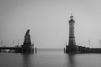 A black and white image features a waterfront scene, including a prominent lighthouse on the right and a large lion sculpture on the left, both on separate platforms extending into the water. Several people can be seen on the platform, and there are flags and boats present, contributing to a serene atmosphere.