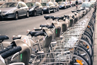 A row of rental bicycles with baskets is lined up on a city street. There are parked cars on the side of the street behind the bicycles, and buildings are visible in the background. Some people can be seen in the distance.