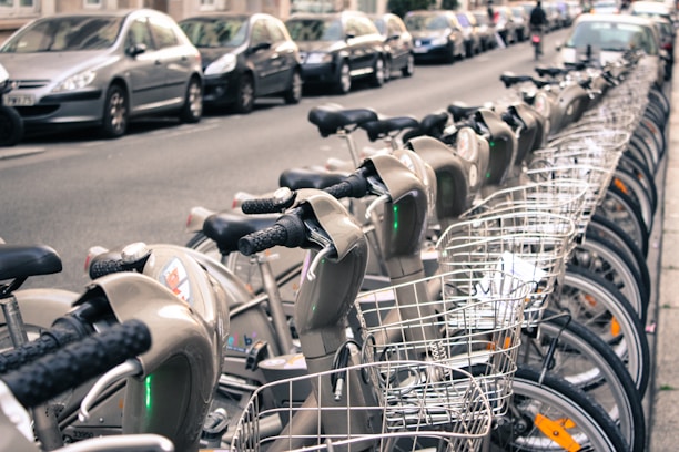 A row of rental bicycles with baskets is lined up on a city street. There are parked cars on the side of the street behind the bicycles, and buildings are visible in the background. Some people can be seen in the distance.