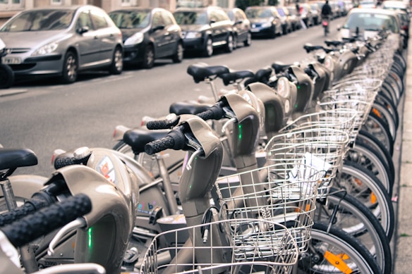 A row of rental bicycles with baskets is lined up on a city street. There are parked cars on the side of the street behind the bicycles, and buildings are visible in the background. Some people can be seen in the distance.