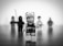 A close-up black-and-white photograph of a distressed-looking LEGO figurine with a worried expression, standing in focus on a smooth surface. In the background, there are four out-of-focus LEGO figurines arranged in a line.
