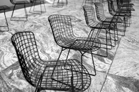 A series of wireframe chairs organized in rows on a marbled stone floor. The chairs have a sleek, modern design with grid-like metal construction. The image is in black and white, enhancing the contrast between the chairs and the textured floor.