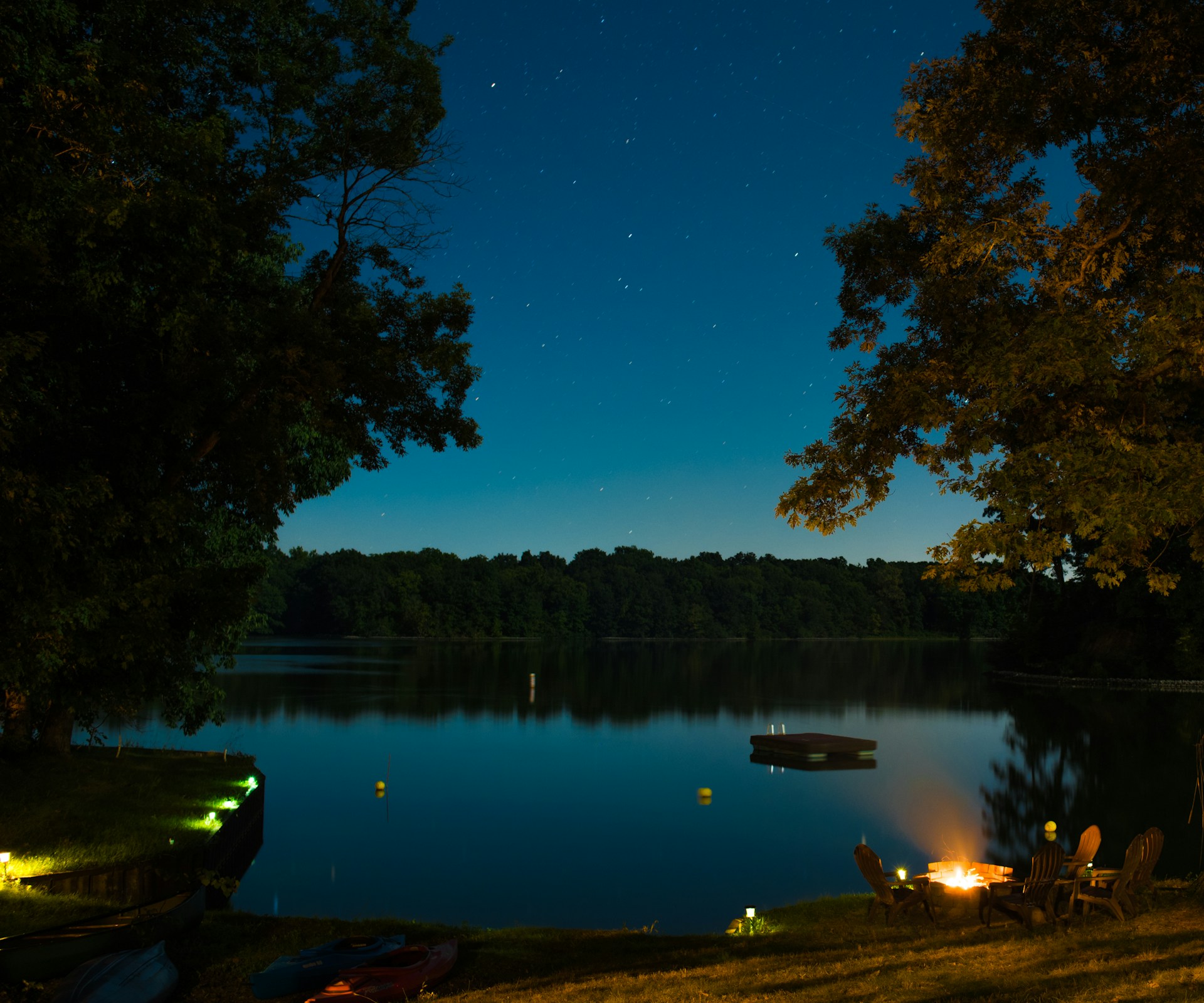 A serene lakeside campsite under a starry night sky, with a cozy fire flickering and tents nestled among tall pines.