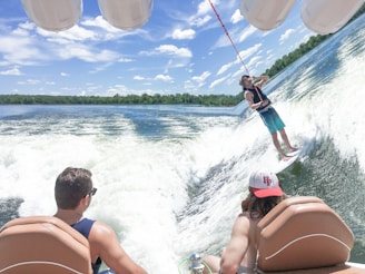 A person is wakeboarding on a large wave behind a boat. Two people are observing from inside the boat, seated and wearing casual attire. The water is clear with visible splashes, and the sky is bright with scattered clouds.