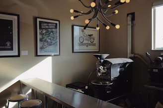 A modern coffee roastery setup includes a commercial coffee roasting machine, a stainless steel workbench with stools, and framed vintage motorcycle posters on the walls. An industrial-looking light fixture with numerous exposed bulbs hangs from the ceiling, adding to the stylish decor. The room is lit by natural light coming from a window on the right.
