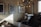 A modern coffee roastery setup includes a commercial coffee roasting machine, a stainless steel workbench with stools, and framed vintage motorcycle posters on the walls. An industrial-looking light fixture with numerous exposed bulbs hangs from the ceiling, adding to the stylish decor. The room is lit by natural light coming from a window on the right.