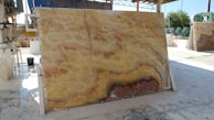 A large, polished stone slab with a distinctive pattern featuring swirling hues of yellow, orange, and brown is propped up vertically in an industrial setting. Various machinery and equipment are visible in the background, along with a partially covered area providing shade. The ground appears to be wet, reflecting the industrial processes that take place here.