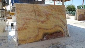 A large, polished stone slab with a distinctive pattern featuring swirling hues of yellow, orange, and brown is propped up vertically in an industrial setting. Various machinery and equipment are visible in the background, along with a partially covered area providing shade. The ground appears to be wet, reflecting the industrial processes that take place here.