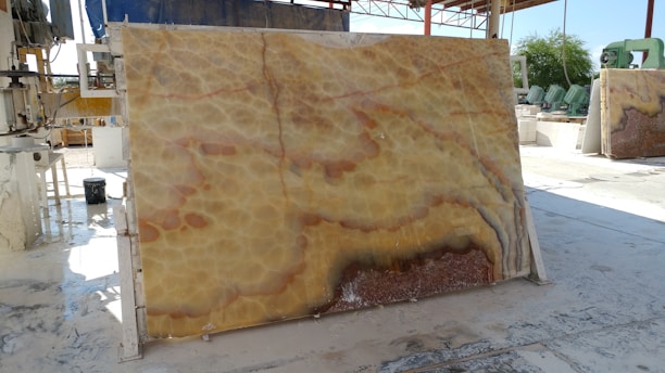 A large, polished stone slab with a distinctive pattern featuring swirling hues of yellow, orange, and brown is propped up vertically in an industrial setting. Various machinery and equipment are visible in the background, along with a partially covered area providing shade. The ground appears to be wet, reflecting the industrial processes that take place here.