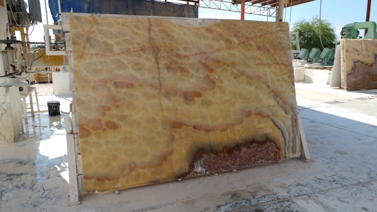 A large, polished stone slab with a distinctive pattern featuring swirling hues of yellow, orange, and brown is propped up vertically in an industrial setting. Various machinery and equipment are visible in the background, along with a partially covered area providing shade. The ground appears to be wet, reflecting the industrial processes that take place here.