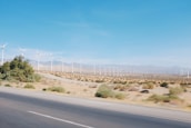 A wide expanse of desert landscape with a row of wind turbines stretching into the distance. The turbines are evenly spaced and surrounded by sparse vegetation. The sky is clear and blue, providing a stark contrast to the brown earth.
