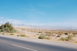 A wide expanse of desert landscape with a row of wind turbines stretching into the distance. The turbines are evenly spaced and surrounded by sparse vegetation. The sky is clear and blue, providing a stark contrast to the brown earth.