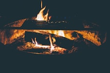 A cozy campfire with flames dancing on wooden logs, casting a warm glow in a low-light setting. The grill above the fire hints at a potential for outdoor cooking or gathering.