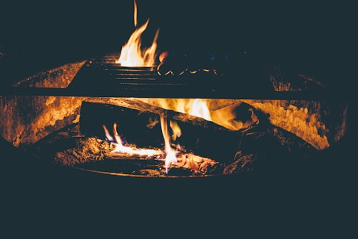 A cozy campfire with flames dancing on wooden logs, casting a warm glow in a low-light setting. The grill above the fire hints at a potential for outdoor cooking or gathering.