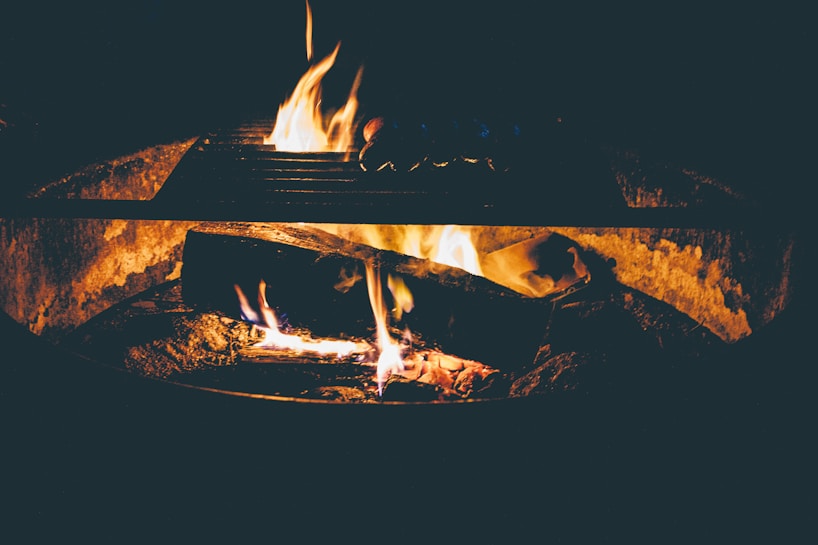 A cozy campfire with flames dancing on wooden logs, casting a warm glow in a low-light setting. The grill above the fire hints at a potential for outdoor cooking or gathering.