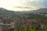 A residential area nestled within a lush, hilly landscape. The foreground features several modern houses with red and brown roofs, surrounded by greenery. In the background, hills and trees stretch under a cloudy yet bright sky, indicating a serene, late afternoon ambiance.