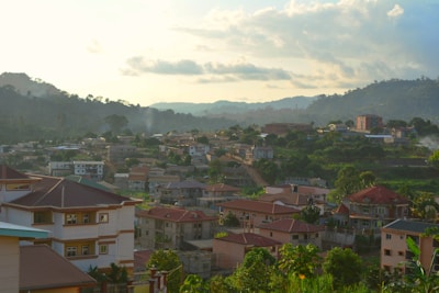 A residential area nestled within a lush, hilly landscape. The foreground features several modern houses with red and brown roofs, surrounded by greenery. In the background, hills and trees stretch under a cloudy yet bright sky, indicating a serene, late afternoon ambiance.