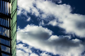 A modern glass building is partially visible on the left, featuring a grid-like pattern of metal or dark lines. Large, fluffy white clouds are scattered across a deep blue sky, creating a contrast between the man-made structure and the natural elements.