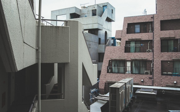 Concrete and brick buildings with geometric shapes form an urban landscape. Several windows, balconies, and air conditioning units are visible. The architecture is modern and somewhat industrial.