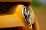 Close-up of a car's front grill featuring the Toyota logo, with the surrounding area predominantly yellow and a blurred background.