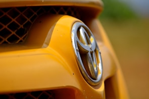 Close-up of a car's front grill featuring the Toyota logo, with the surrounding area predominantly yellow and a blurred background.