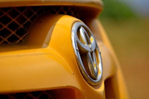Close-up of a car's front grill featuring the Toyota logo, with the surrounding area predominantly yellow and a blurred background.